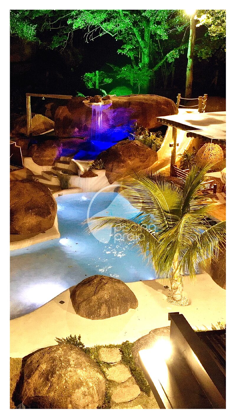 Piscina noturna entre rochas, com iluminação azul e deck de madeira