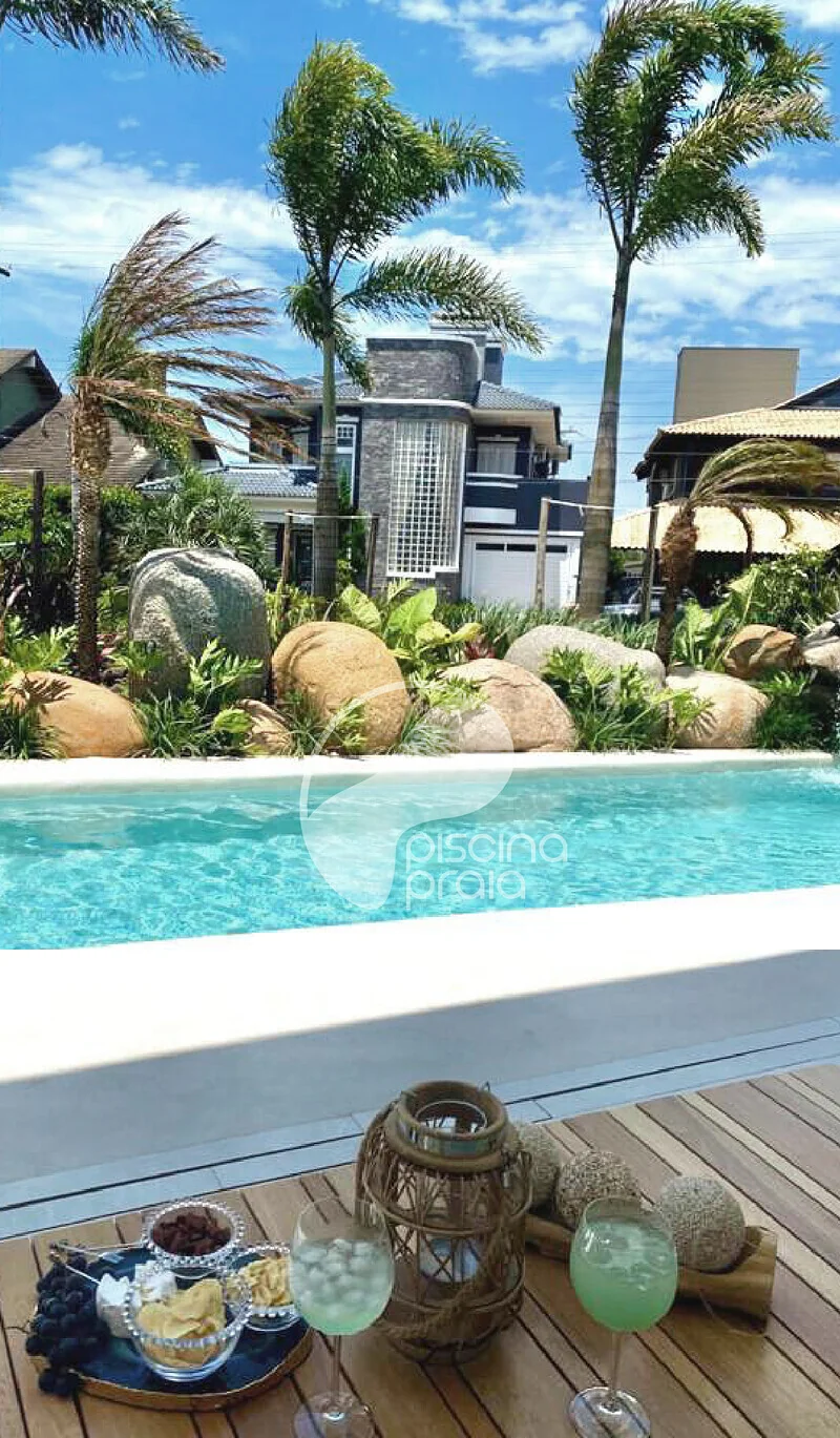 Piscina de condomínio com coqueiros e pedras, e drinks servidos no deck
