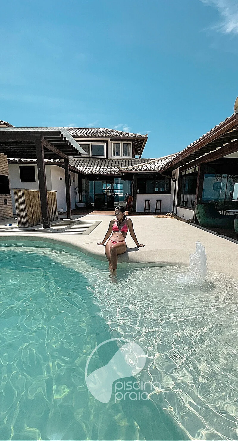 Piscina com prainha de areia em frente à casa, com pessoa sentada na borda