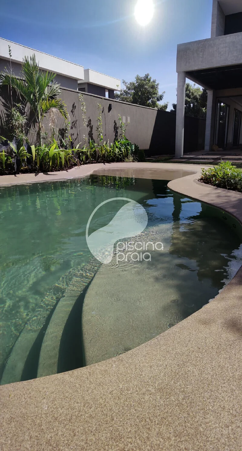 Piscina de fundo escuro com prainha de areia junto à casa de concreto