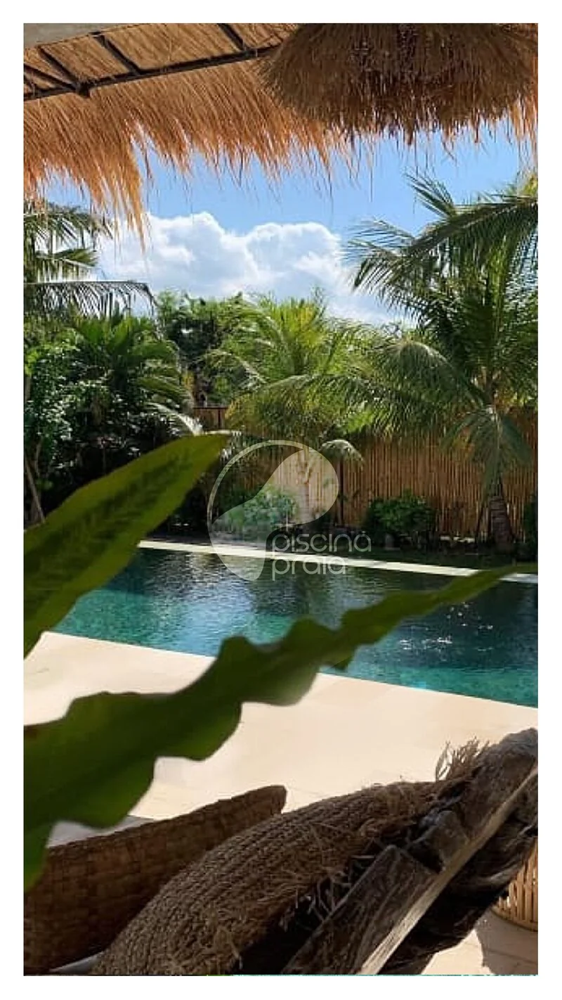 Piscina vista da sombra de um quiosque de palha, com coqueiros ao redor