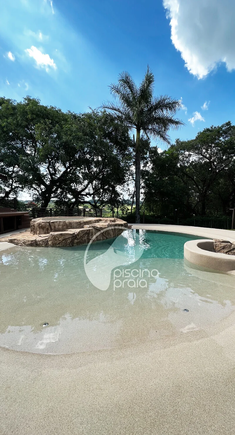 Piscina com prainha larga de areia, rochas e árvores ao fundo